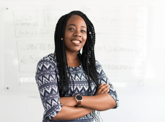 Smiling woman with braids and a professional attire standing confidently in front of a whiteboard.