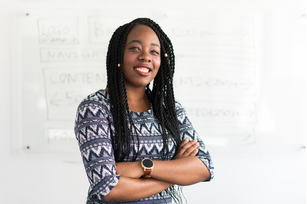 Smiling woman with braids and a professional attire standing confidently in front of a whiteboard.