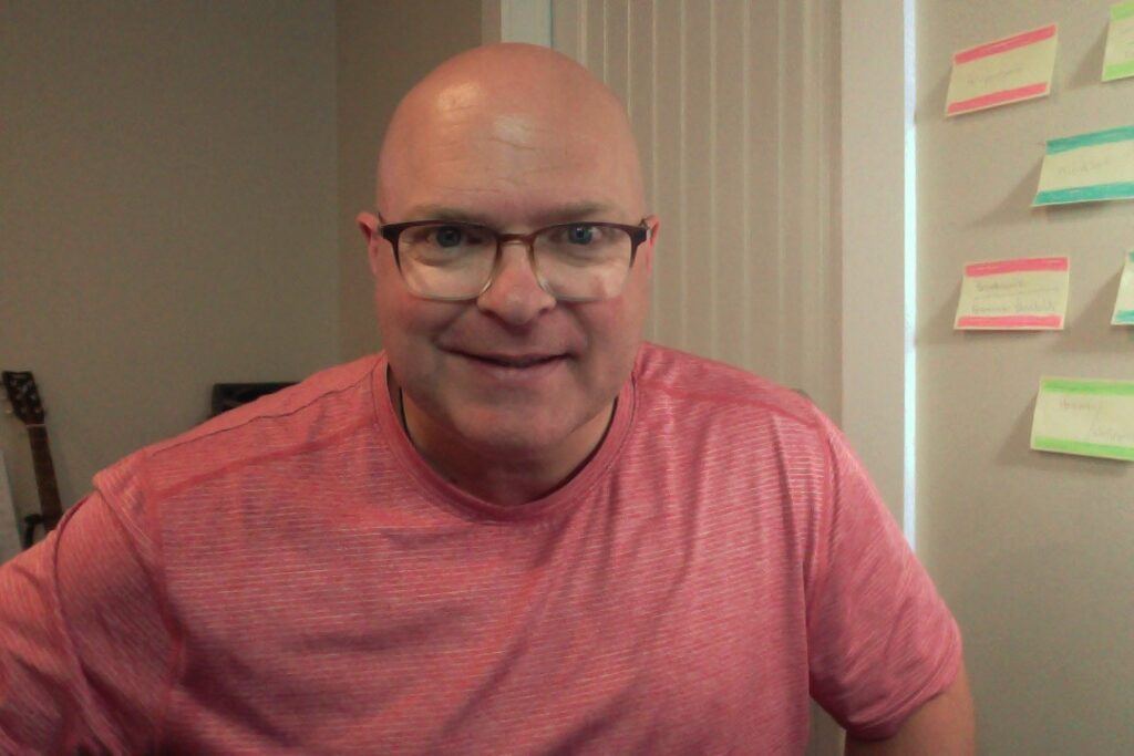 Smiling bald man with glasses wearing a pink t-shirt.