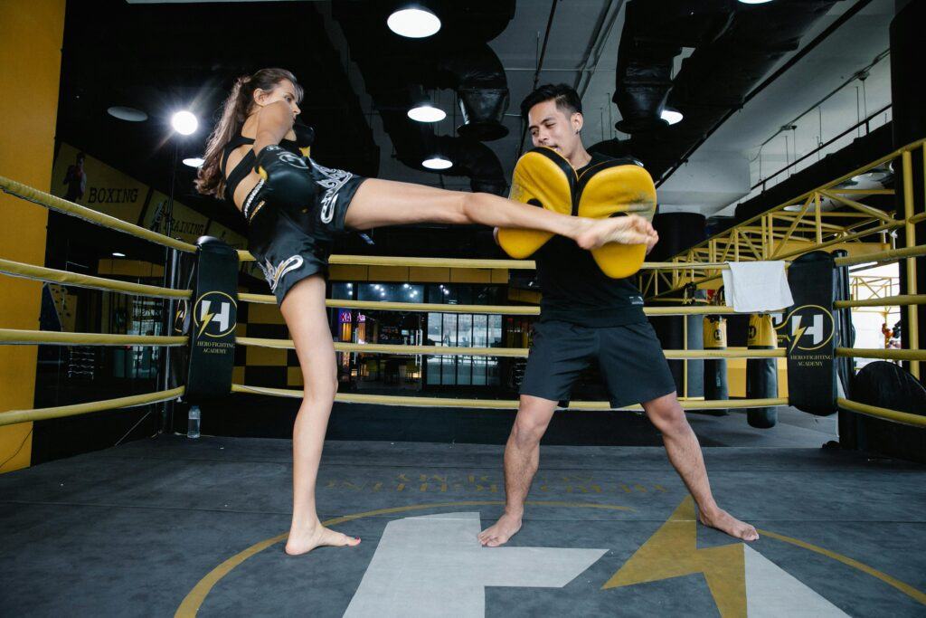 Female Muay Thai fighter practicing high kick with male coach holding training pads in a boxing gym.