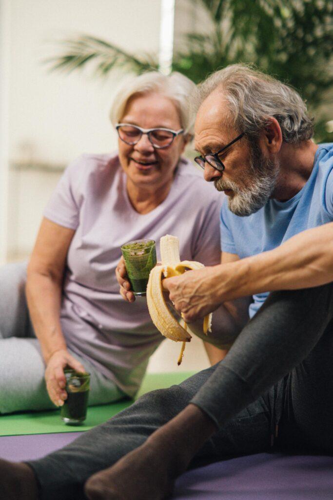 An older couple sharing a light-hearted moment with healthy snacks on yoga mats.