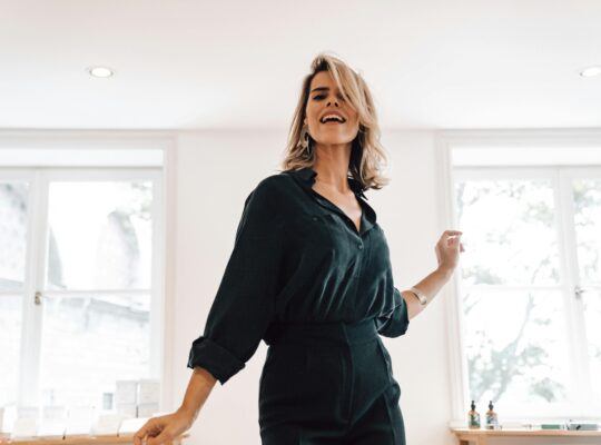 Confident, smiling woman in a black jumpsuit standing in a bright, modern office setting.