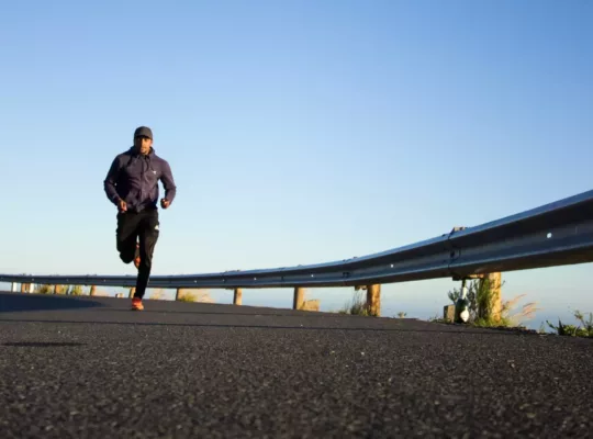 A black man in running attire strides towards the camera on a curved elevated roadway with a guardrail.