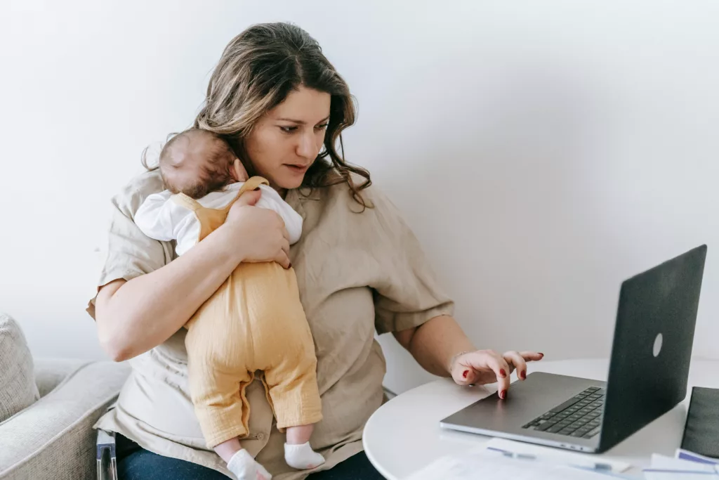 Busy mom holding a baby while working on a laptop, symbolizing multitasking and trying to balance parenting and personal goals.